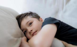 filmische portret gelukkige jonge jongen liggend op de bank uitkijkend met een lachend gezicht, positief kind rustend in de woonkamer met fel licht schijnt op de lente of zomer. kind ontspannen tv kijken in het weekend foto