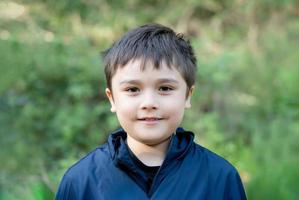 buitenshuis portret knappe jonge jongen die naar camera kijkt met een lachend gezicht, gezond kind met een blij gezicht dat alleen in het park staat, positief kind dat buiten speelt in de lente of zomervakantie foto