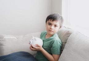 gelukkige jongen met spaarvarken met lachend gezicht. indoor portret van een vrolijk kind met geldbesparende box.schooljongen die financiële verantwoordelijkheid leert en plannen maakt over sparen voor toekomstig concept foto