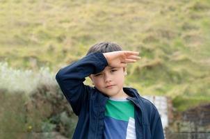 portret van jonge jongen met hand omhoog die fel zonlicht afschermt dat in zijn ogen schijnt, kind dat in groene velden staat, vrolijk kind dat plezier heeft met buiten spelen in de lente of zomer foto