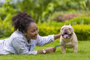 Afro-Amerikaanse vrouw speelt met haar Franse bulldog-puppy terwijl ze in het grasveld ligt na 's ochtends te hebben geoefend in het park foto