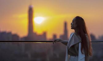 vrouw die kijkt en geniet van het uitzicht op de zonsondergang vanaf het balkon met de ondergaande zon achter de wolkenkrabber in het drukke stedelijke centrum met eenzaamheid voor eenzaamheid, eenzaamheid en dromen van vrijheidslevensstijl foto