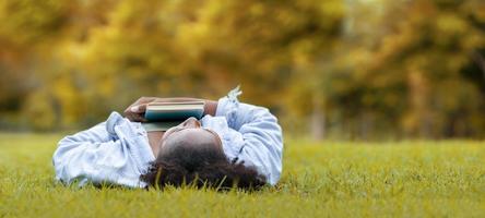 Afro-Amerikaanse vrouw ligt in het grasveld in het openbare park met boek in haar hand in de herfst voor herfstseizoen en en onderwijsconcept met kopieerruimte foto
