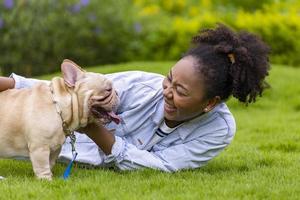 Afro-Amerikaanse vrouw speelt met haar Franse bulldog-puppy terwijl ze in het grasveld ligt na 's ochtends te hebben geoefend in het park foto