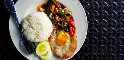 bovenaanzicht van roergebakken basilicum met inktvis, rijst, gesneden komkommer en gebakken ei in thaise stijl op witte plaat met kopieerruimte. plat leggen van beroemd en pittig eten op zwarte roestvrijstalen achtergrond. foto