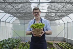 een blanke mannelijke boer kijkt en geeft een mand met verse groenten aan de camera met een gelukkige glimlach in de plantagekas. tuinman man verzamelt natuurlijke biologische producten van landbouwkwekerijgewassen. foto