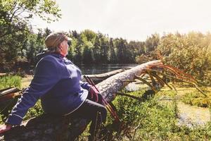 oudere vrouw zit op een boomtak aan de oever van het meer en geniet van een zomerdag in de buitenlucht. textuur en natuur welzijn concept achtergrond foto