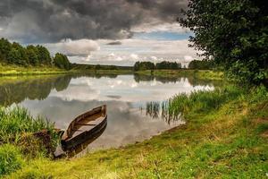 oude houten boot aan de oever van een brede rivier in bewolkte dag voor de storm foto