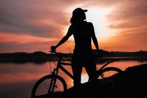 zomer silhouet landschap. meisje met een fiets bij het meer tegen de achtergrond van de ondergaande zon, de karmozijnrode zonsondergang foto