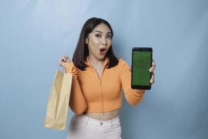 Portret van een Aziatische mooie jonge vrouw die verrast staat met een online boodschappentas en haar smartphone laat zien, studio-opname geïsoleerd op blauwe achtergrond foto