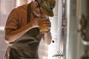 een jonge man werkt in een brouwerij en controleert de kwaliteit van ambachtelijk bier. de brouwerij-eigenaar proeft de beste bieren uit bach. de shortcut van een man vult een glas bier met foto