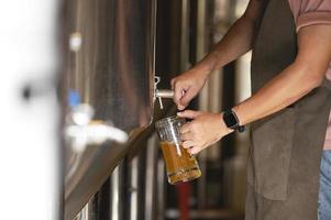 een jonge man werkt in een brouwerij en controleert de kwaliteit van ambachtelijk bier. de brouwerij-eigenaar proeft de beste bieren uit bach. de shortcut van een man vult een glas bier met foto