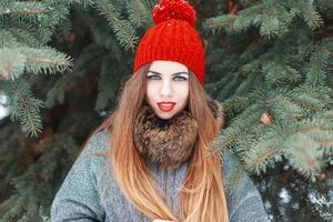 portret van een mooie jonge vrouw met rode lippen en een rode gebreide muts op de achtergrond van een groene spar foto