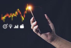 financiële en investeringsconcepten de hand van de man die een smartphone vasthoudt, toont grafieken en pictogrammen die betrouwbaarheid en een analyse van aandelengrafieken tonen. om forex, valutawissel, olie, goud of andere indexen te verhandelen. foto