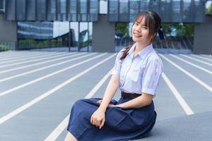 mooie Aziatische middelbare school student meisje in het schooluniform met een glimlach vol vertrouwen terwijl ze gelukkig naar de camera kijkt met het gebouw op de achtergrond. foto