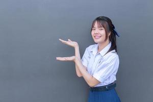 schattig aziatisch middelbare school student meisje in het schooluniform met een zelfverzekerde glimlach terwijl ze naar de camera kijkt om iets vrolijks te presenteren met grijs op de achtergrond. foto