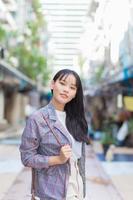 zelfverzekerde professionele jonge Aziatische zakenvrouw die een bruin gestreepte blazer en schoudertas draagt, glimlacht vrolijk en kijkt naar de camera terwijl ze door de oude stad naar haar werk pendelt. foto