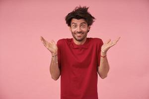 portret van een jonge man met open ogen met wild haar die over een roze achtergrond staat met opgeheven handpalmen, casual kleding draagt, naar de camera kijkt met een brede glimlach en wenkbrauwen optrekt foto