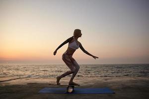 jonge slanke getatoeëerde vrouw met goede fysieke conditie balancerend aan boord met uitzicht op zee, oefeningen doend met balancer aan de kust in de vroege ochtend, sportieve top en leggins dragen foto