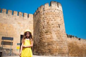 een toeristisch meisje met een gele hoed en een zomerjurk loopt langs de straat van de oude stad, gemaakt van steen met een fort. rondleiding. het kind verdwaalde, op zoek naar ouders foto