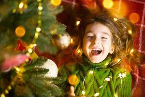 een meisje met lang haar en slingers ligt op een rode plaid onder een kerstboom met speelgoed in een warme gebreide trui. Kerstmis, Nieuwjaar, emoties van kinderen, vreugde, verwachting van een wonder en geschenken foto