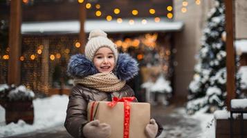 portret van een vrolijk meisje met een geschenkdoos voor kerstmis op een stadsstraat in de winter met sneeuw op een feestelijke markt met versieringen en kerstverlichting. warme kleding, gebreide muts, sjaal en bont. Nieuwjaar foto