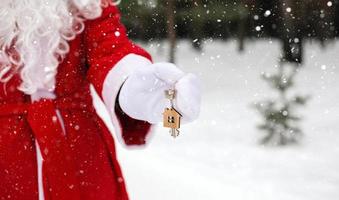 huissleutel met sleutelhanger huisje in handen van de kerstman buiten in de sneeuw. deal voor onroerend goed, aankoop, bouw, verhuizing, hypotheek. gezellig huis. vrolijk kerstfeest, nieuwjaarsboekingsevenement en hal foto