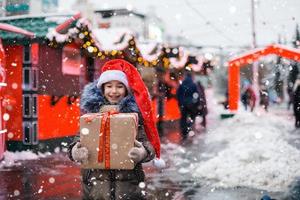 portret van vrolijk meisje in kerstmuts met geschenkdoos voor kerstmis op straat in de winter met sneeuw op feestelijke markt met versieringen en kerstverlichting. warme kleding, gebreide sjaal en bont. Nieuwjaar foto