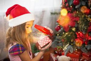 klein meisje in kerstmuts opent een rode doos met een geschenk en een gouden magisch licht in de buurt van de kerstboom. vakantie decor, kerststerren op dennenbomen, nieuwjaar. vreugde, verrassing, de emoties van kinderen. kopieerruimte foto