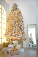 een kerstboom met een gouden decoratie wordt weerspiegeld in de spiegel. nieuwjaar, woondecoratie. groot raam met geel warm licht, comfort in de woonkamer foto
