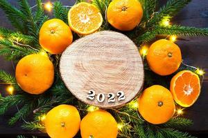 nieuwjaarsvakantie achtergrond op een ronde snede van een boom omringd door mandarijnen, levende dennentakken en gouden lichtslingers, met houten nummers datum 2022. citrusaroma, kerstmis. ruimte voor tekst. foto