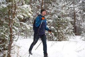 skiër met een rugzak en muts met pompon met skistokken in zijn handen op de achtergrond van een besneeuwd bos. langlaufen in het winterbos, buitensporten, gezonde levensstijl, wintersporttoerisme. foto