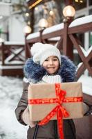 portret van een vrolijk meisje met een geschenkdoos voor kerstmis op een stadsstraat in de winter met sneeuw op een feestelijke markt met versieringen en kerstverlichting. warme kleding, gebreide muts, sjaal en bont. Nieuwjaar foto