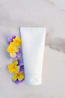 witte plastic crème tube en heldere viooltjesbloemen op marmer, close-up, mockup. huidverzorging schoonheidsproduct, crème of lotion. exotische natuurlijke cosmetica, minimale samenstelling foto