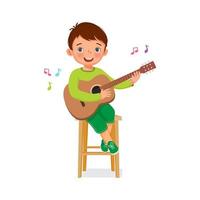 schattige kleine jongen die een ukelele of gitaar speelt terwijl hij op een houten stoel zingt