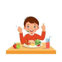 schattige kleine jongen die ontbijt eet met brood, gebakken ei, broccoli, groenten met vork met worst die duim omhoog gebaar toont