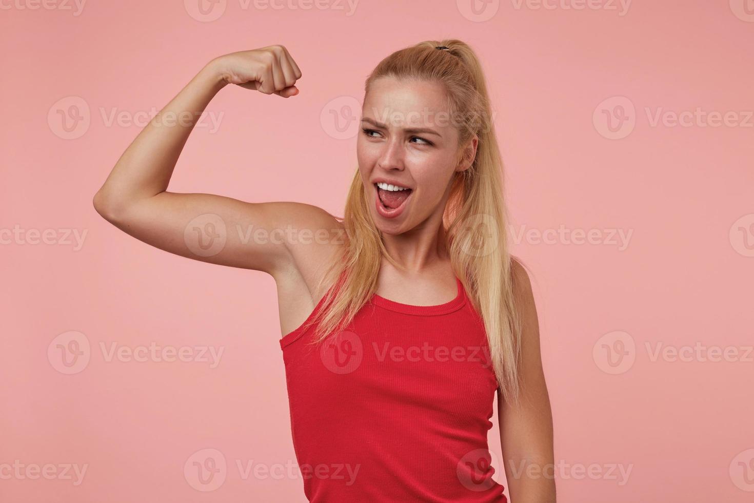 portrait d'une belle jeune femme aux cheveux longs dans des vêtements décontractés montrant son pouvoir avec la main levée, fière de ses biceps, isolée sur fond rose photo