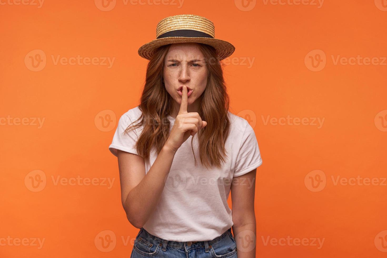 belle jeune femme aux cheveux rouges dans des vêtements décontractés à la recherche d'appareil photo avec l'index levé à sa bouche, faisant un geste de silence sur fond orange