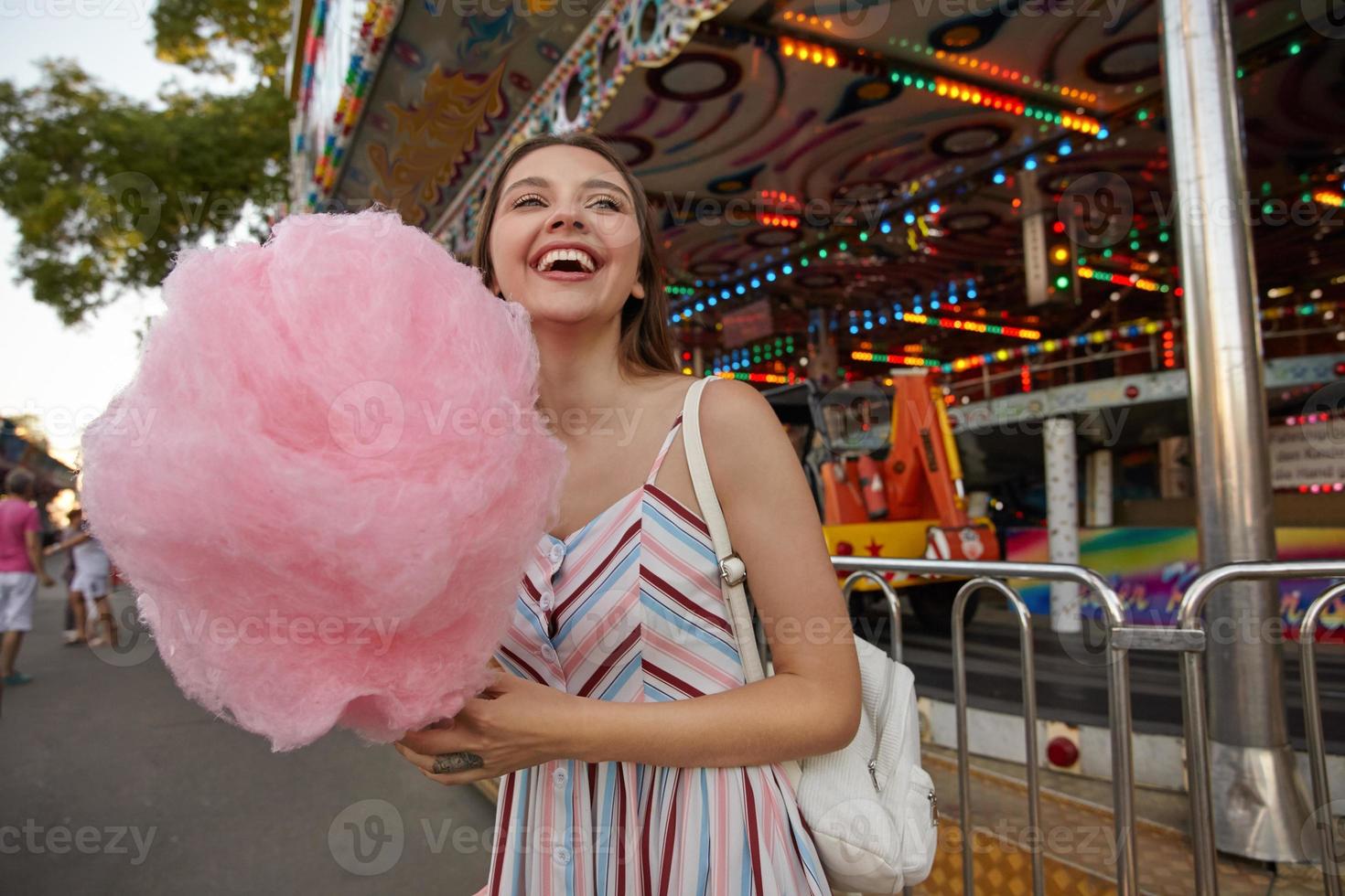 joyeuse jeune femme brune séduisante aux cheveux longs marchant dans un parc d'attractions, vêtue d'une robe d'été et tenant une barbe à papa sur un bâton, regardant de côté et riant joyeusement photo