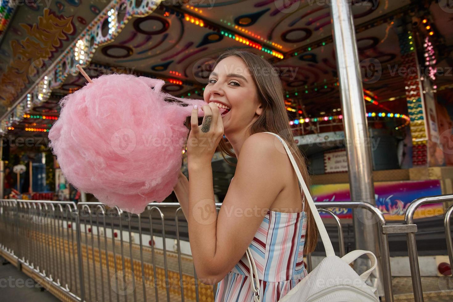 portrait d'une jolie jeune femme joyeuse avec une coiffure décontractée en robe d'été romantique mangeant de la barbe à papa tout en marchant dans un parc d'attractions par temps chaud, concept d'émotions positives photo