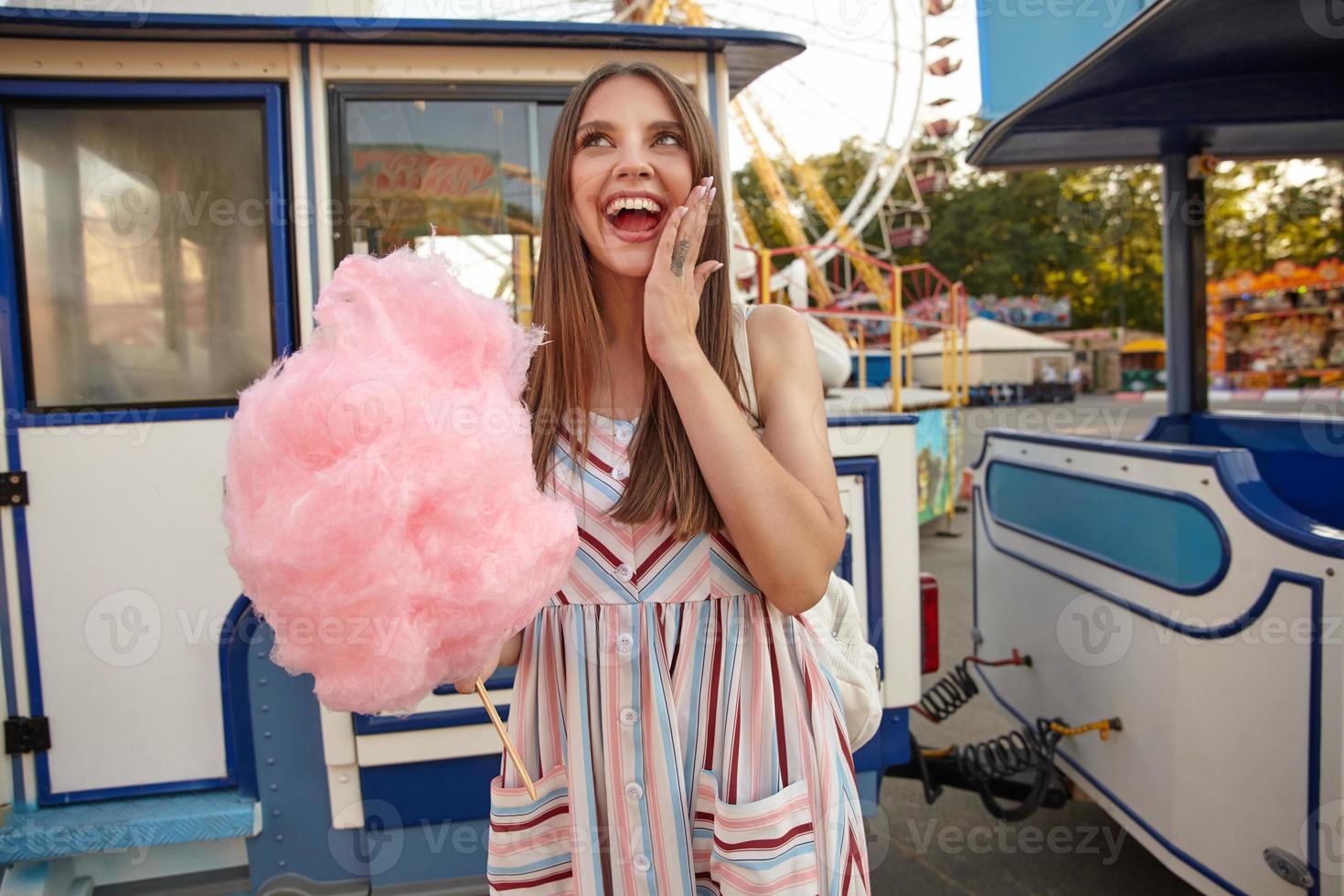 heureuse jeune brune séduisante aux cheveux longs portant une robe d'été à bretelles, posant sur un parc d'attractions avec de la barbe à papa rose à la main, gardant la paume sur sa joue et souriant joyeusement photo