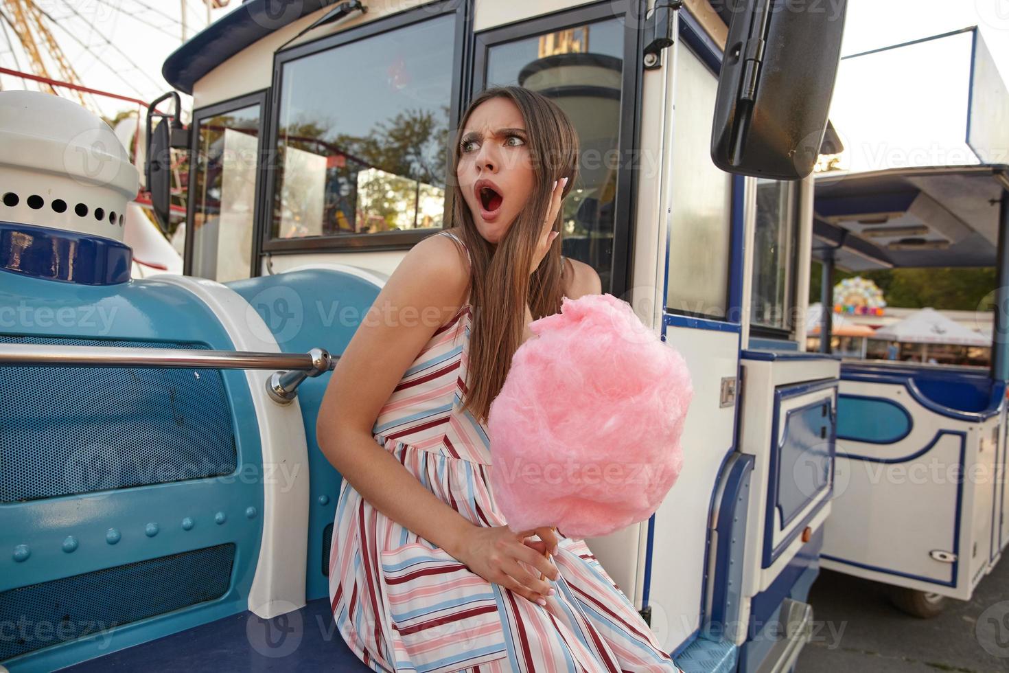 surprise séduisante jeune femme aux cheveux longs avec de la barbe à papa à la main assise au-dessus d'un parc d'attractions par une chaude journée d'été, regardant étonnamment de côté avec la bouche large ouverte et le visage froncé photo