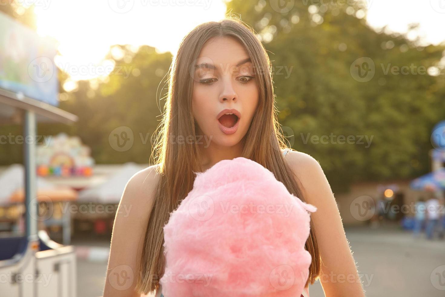 photo en plein air d'une belle jeune femme brune aux cheveux longs posant sur un parc verdoyant par une chaude journée ensoleillée, regardant la barbe à papa rose avec un visage surpris et ouvrant largement la bouche