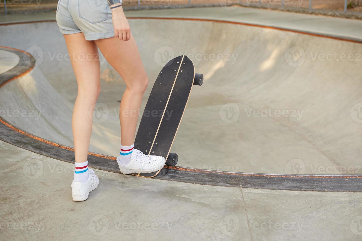 photo en plein air recadrée d'une femme mince en short en jean posant sur une planche à roulettes par temps chaud d'été, va monter à bord dans un parc de cascades