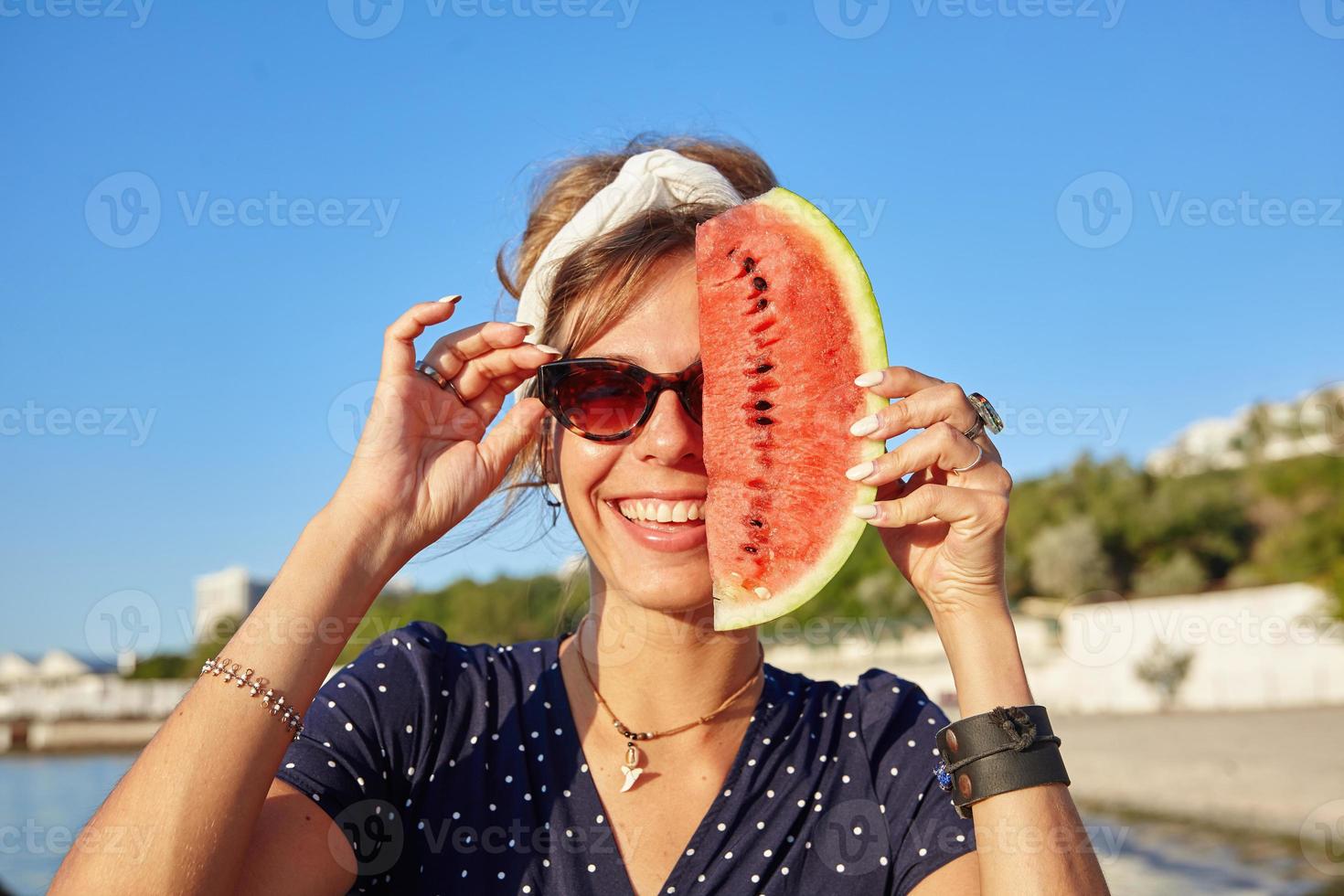 heureuse jeune femme brune en bandeau et lunettes de soleil vintage regardant joyeusement la caméra et tenant une tranche de pastèque par son visage, debout sur la vue sur la mer photo