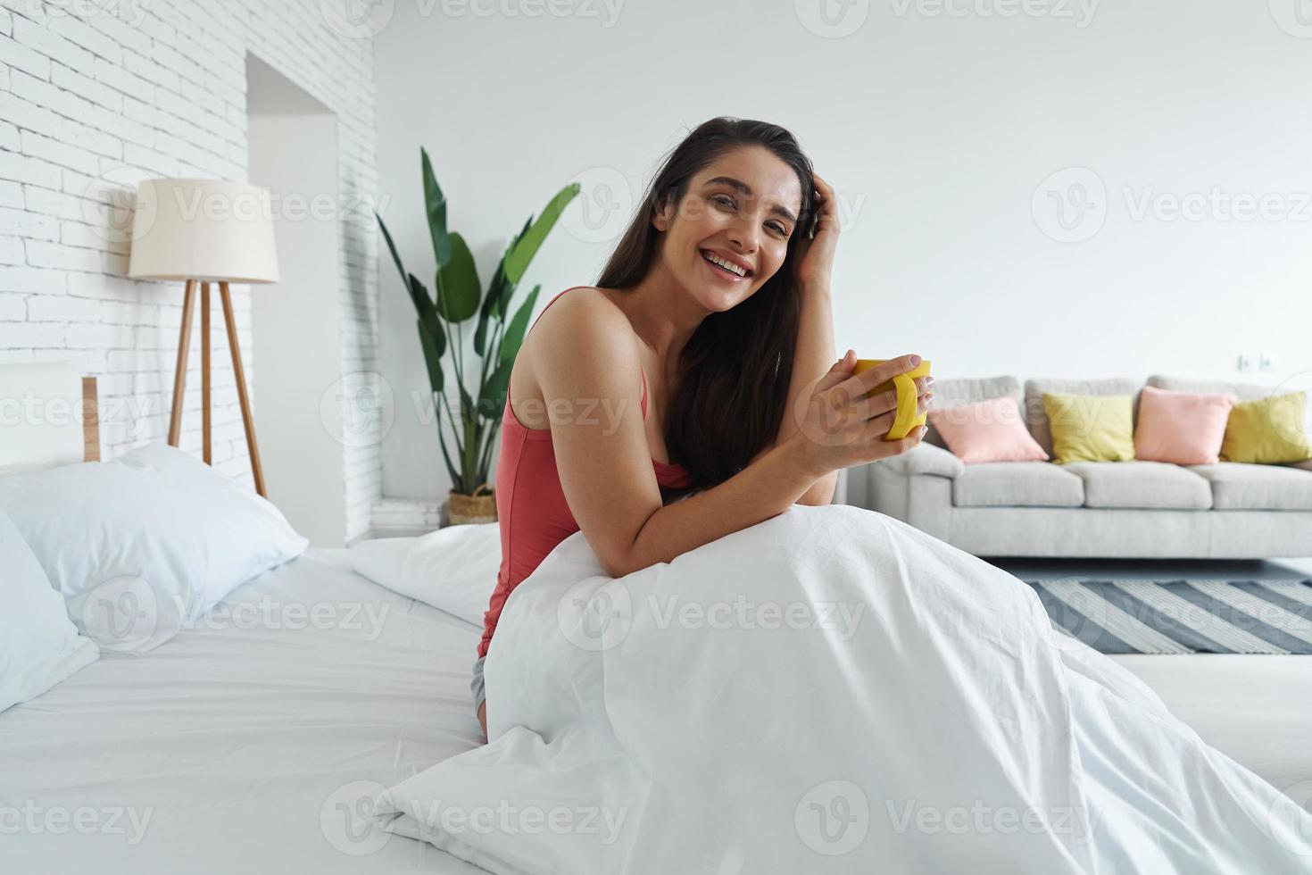 heureuse jeune femme tenant une tasse de café assise dans son lit à la maison photo