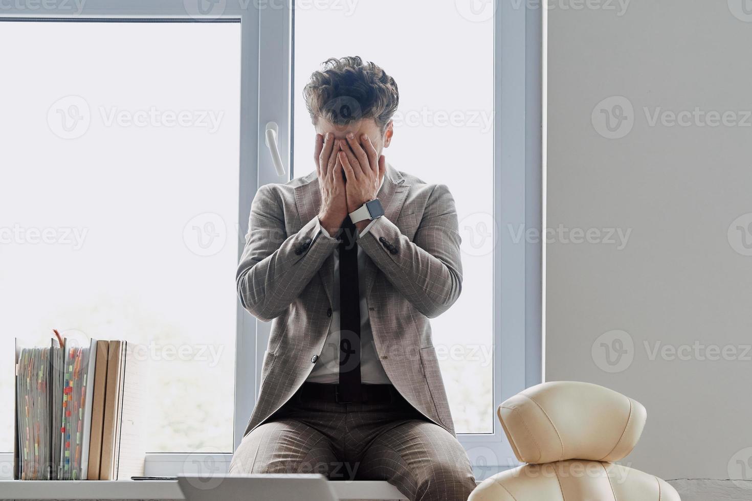jeune homme déprimé couvrant le visage avec les mains tout en étant assis sur le rebord de la fenêtre au bureau photo