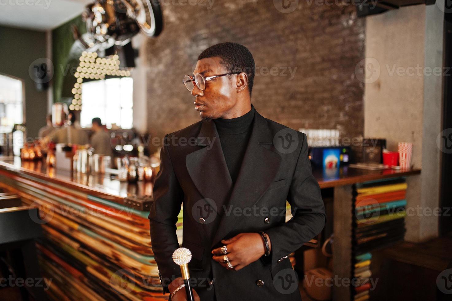 élégant gentleman afro-américain dans une élégante veste noire et des lunettes, tenant un bâton de marche rétro comme un flacon de canne ou une canne basculante avec une poignée en boule de diamant doré. riche homme afro à la mode. photo