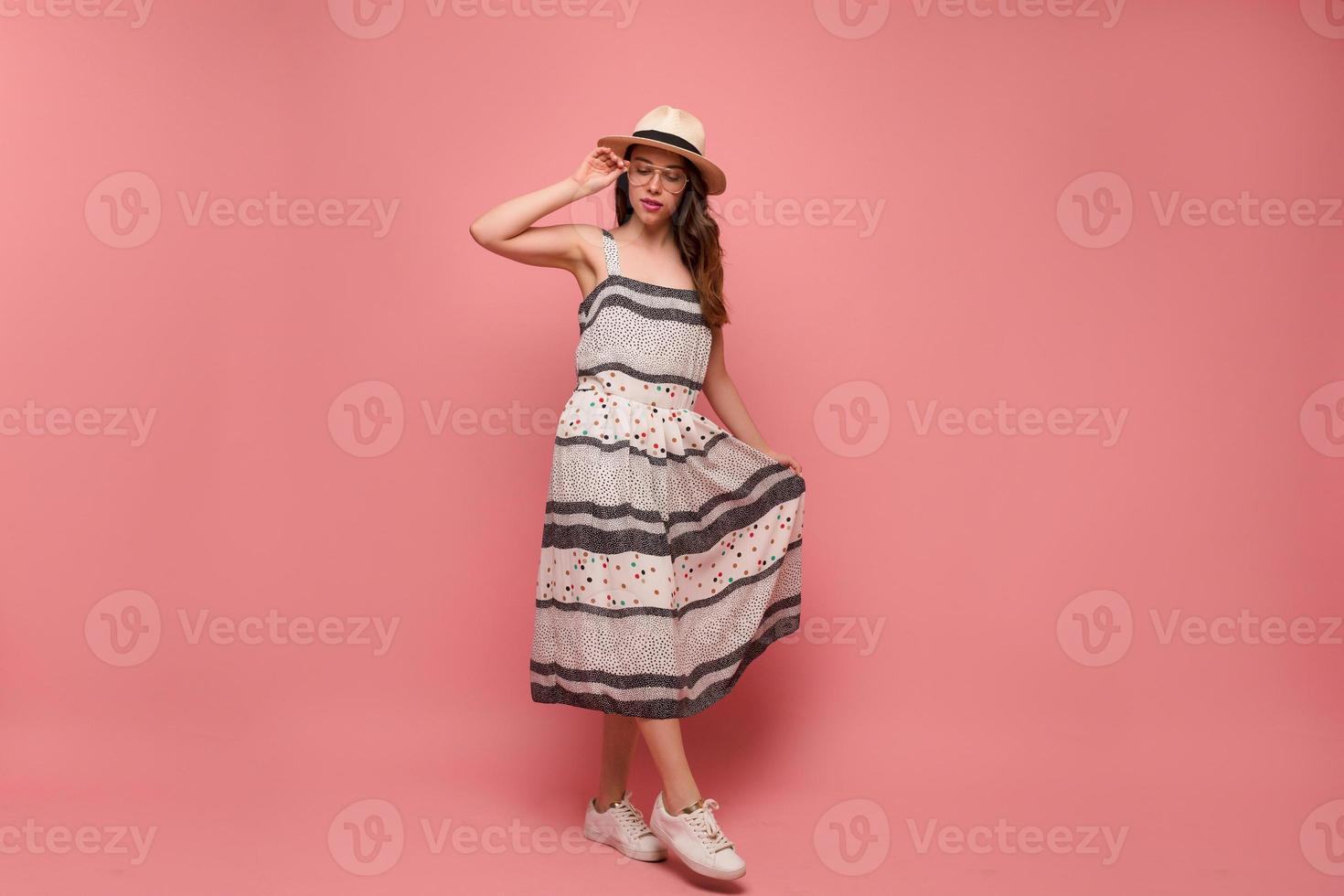 portrait en pied d'une adorable femme élégante en chapeau et robe d'été touche le chapeau et pose sur fond rose photo