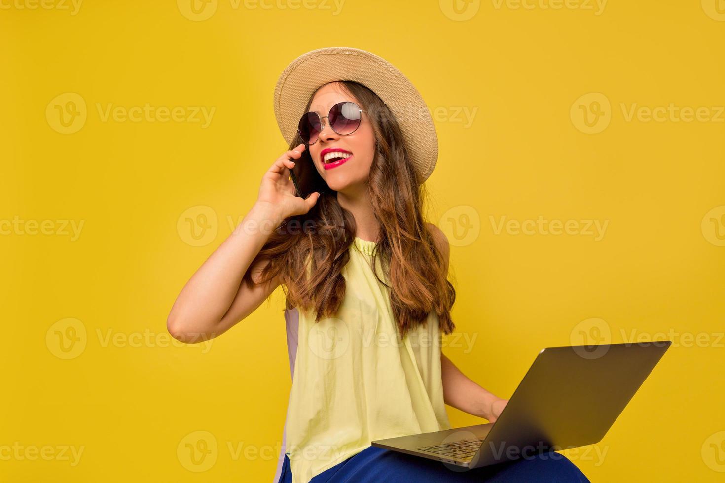 charmante jolie femme portant un chapeau d'été et des lunettes parlant au téléphone et travaillant avec un ordinateur portable sur fond jaune isolé avec un sourire adorable, de vraies émotions heureuses, des vacances photo